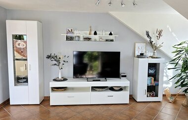 Wohnzimmer mit weißem TV-Schrank, großem Flachbildfernseher, zwei weißen Vitrinenschränken, Regalen mit Dekoration und Pflanzen auf braunem Fliesenboden | © Familie Fuhrmann