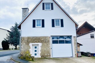 Weißes Haus mit steilem Dach, drei Fenstern mit schwarzen Fensterläden, einer weißen Tür und einer großen weißen Garagentür, im Hintergrund ein Glockenturm | © Ferienwohnung Etzl