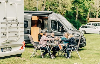 Ein weisses und ein graues Wohnmobil. 2 Männer und 2 Frauen die an einem Campingtisch sitzen und sich zuprosten. Buchen im Odenwald | © Touristikgemeinschaft Odenwald e.V.