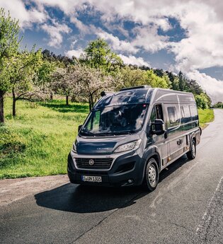 Dunkler Kastenwagen fährt auf einer Straße. Links vom Wohnmobil ist eine Streuobstwiese.| Odenwald | © Touristikgemeinschaft Odenwald e.V.
