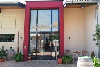 Blick auf den Eingangsbereich mit Glasfront vom Weingut Hofäcker in Weikersheim-Queckbronn. Im Hof davor ist ein Sitzgruppe sowie Blumen und Weinfässer als Dekoration zu sehen. | © Touristinfo Weikersheim