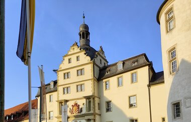 Eingang zum Residenzschloss Mergentheim bei Sonnenuntergang. Im Vordergrund sind rosa Blumen. | © @reishonger