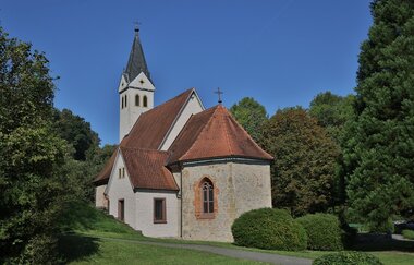 Die Kirche hat einen spitzen Turm und ist von grünen Bäumen umgeben | © Touristikgemeinschaft Hohenlohe e. V. | Andi Schmid