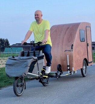 Ein Mann mit Jeans und gelbem T-Shirt bekleidet fährt auf einer asphaltierten Strasse mit einem Lastenfahrrad an dem ein kleiner Fahrradwohnwagen aus Holz angehängt ist | © Dennis Hirschmann