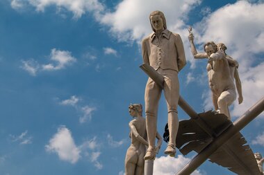 Am Ortseingang von Lauffen am Neckar erhebt sich das eindrucksvolle Kunstwerk „Hölderlin im Kreisverkehr“ des Bildhauers Peter Lenk. Auf einem geschwungenen „H“ balanciert eine überdimensionale Schreibfeder, auf der Friedrich Hölderlin als Kind und als ju | © Stadt Lauffen am Neckar